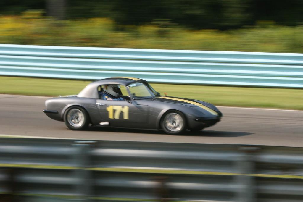 Ein Auto mit einem Helmträger fährt auf einer von Barrieren gesäumten Straße, mit Bäumen im Hintergrund.