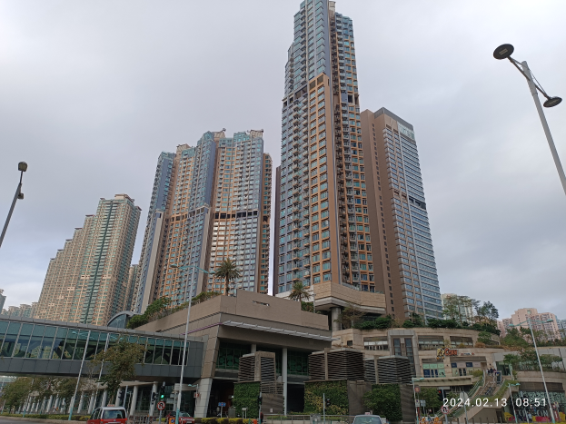 Eine Stadtstraße mit hohen Gebäuden, Fahrzeugen, Laternenpfählen, einer Brücke, Bäumen, Pflanzen und einem bewölkten Himmel, mit einem prominenten Wohnhaus im Vordergrund.