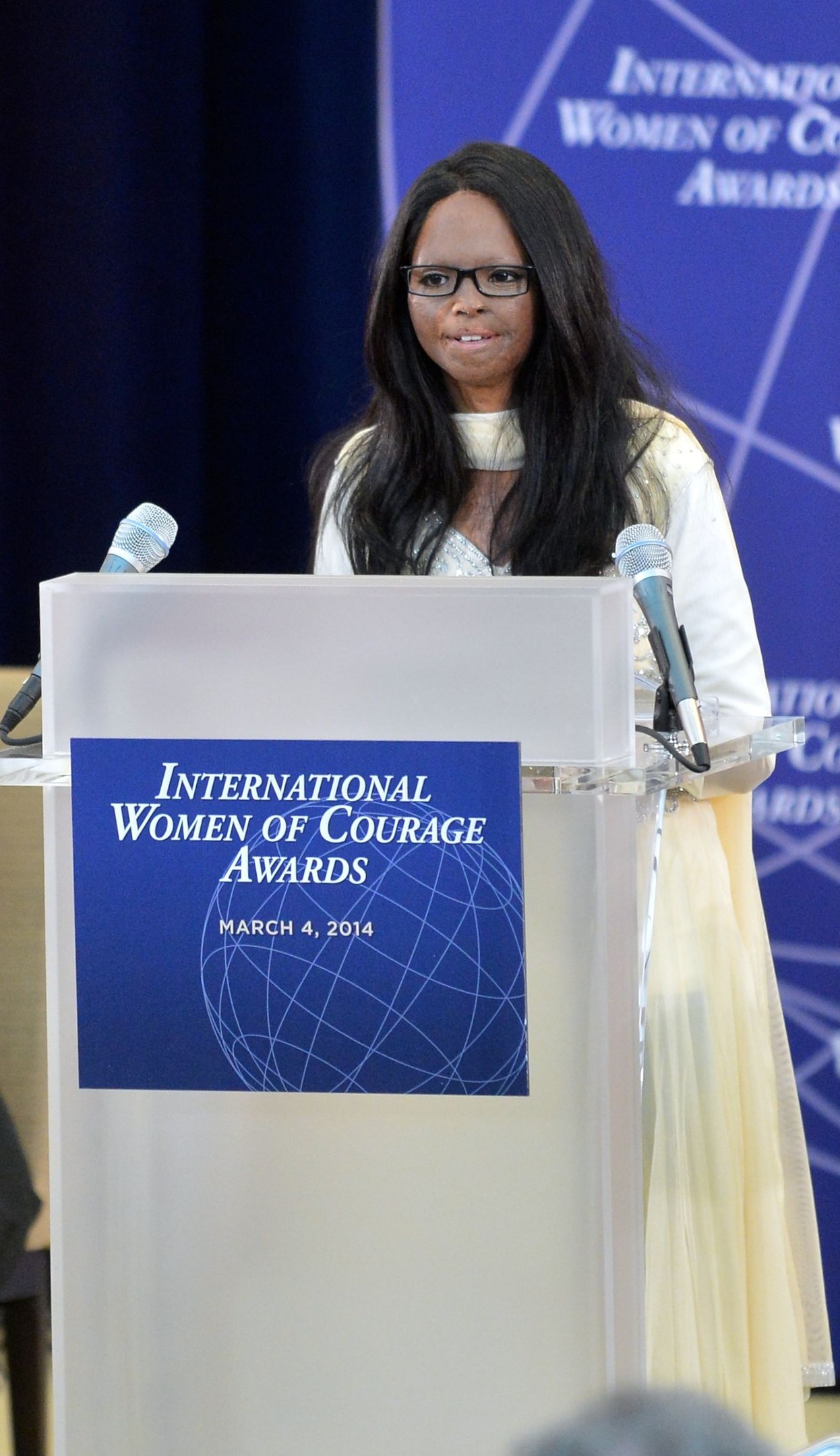 Woman in a white dress and spectacles standing at a podium with two microphones and a board, speaking at the "International Women of Courage Awards 2014" event with a blue banner in the background.
