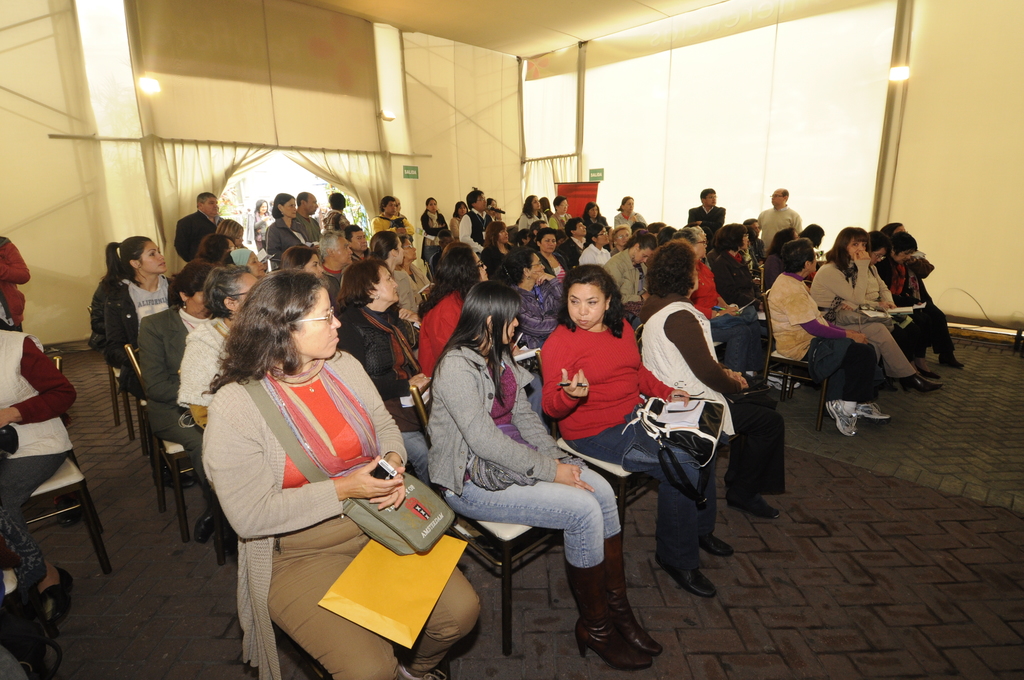 Ein voller Saal mit vielen sitzenden Personen und zwei sichtbaren Ausgängen, durch die einige Menschen eintreten.