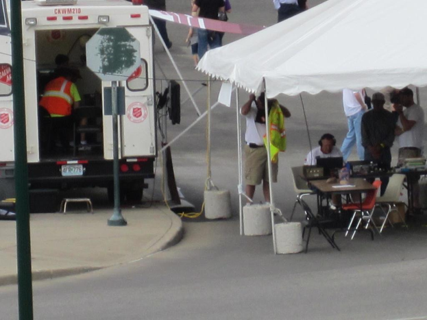 Ein Krankenwagen steht links von einer Stra√üe, w√§hrend auf der rechten Seite ein Zelt viele Menschen sch√ľtzt, mit einem Tisch und einem Stuhl in der N√§he.