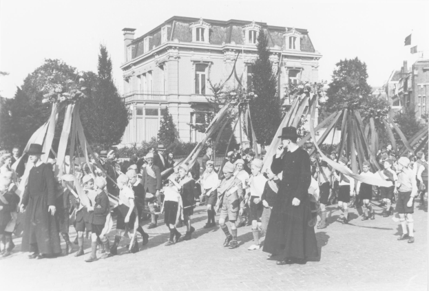 Schwarzes und weißes Bild einer Gruppe von Menschen, die eine Straße entlanggehen und Fähnchen halten, mit Hüten, Maibaum, Bäumen, Gebäuden und Himmel im Hintergrund.