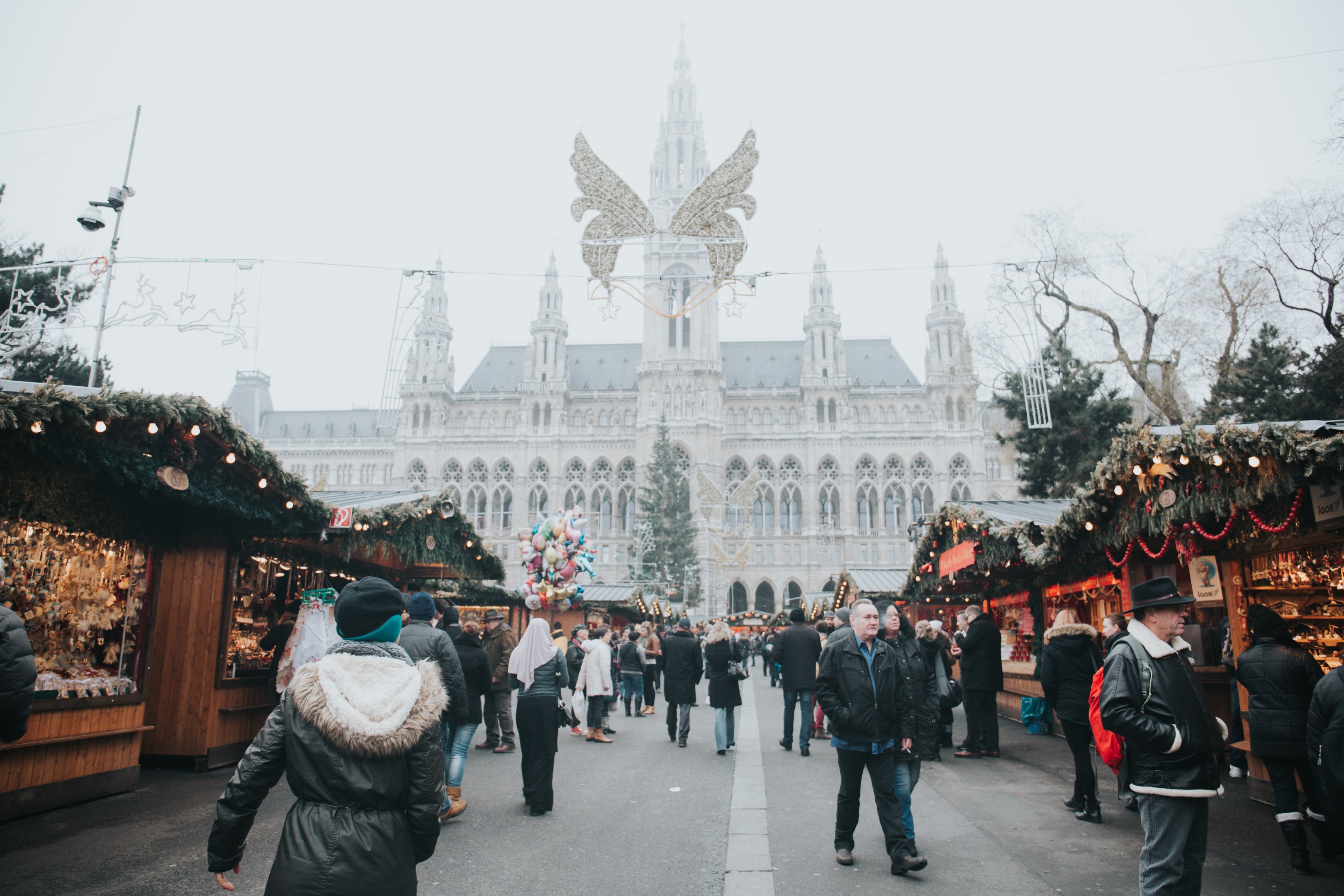 Ein lebendiger Weihnachtsmarkt in Wien, Österreich, mit Menschen, die herumlaufen, Ständen, die mit Lichtern und festlichen Décor geschmückt sind, einem Gebäude mit Fenstern im Hintergrund, Bäumen und einem klaren blauen Himmel.