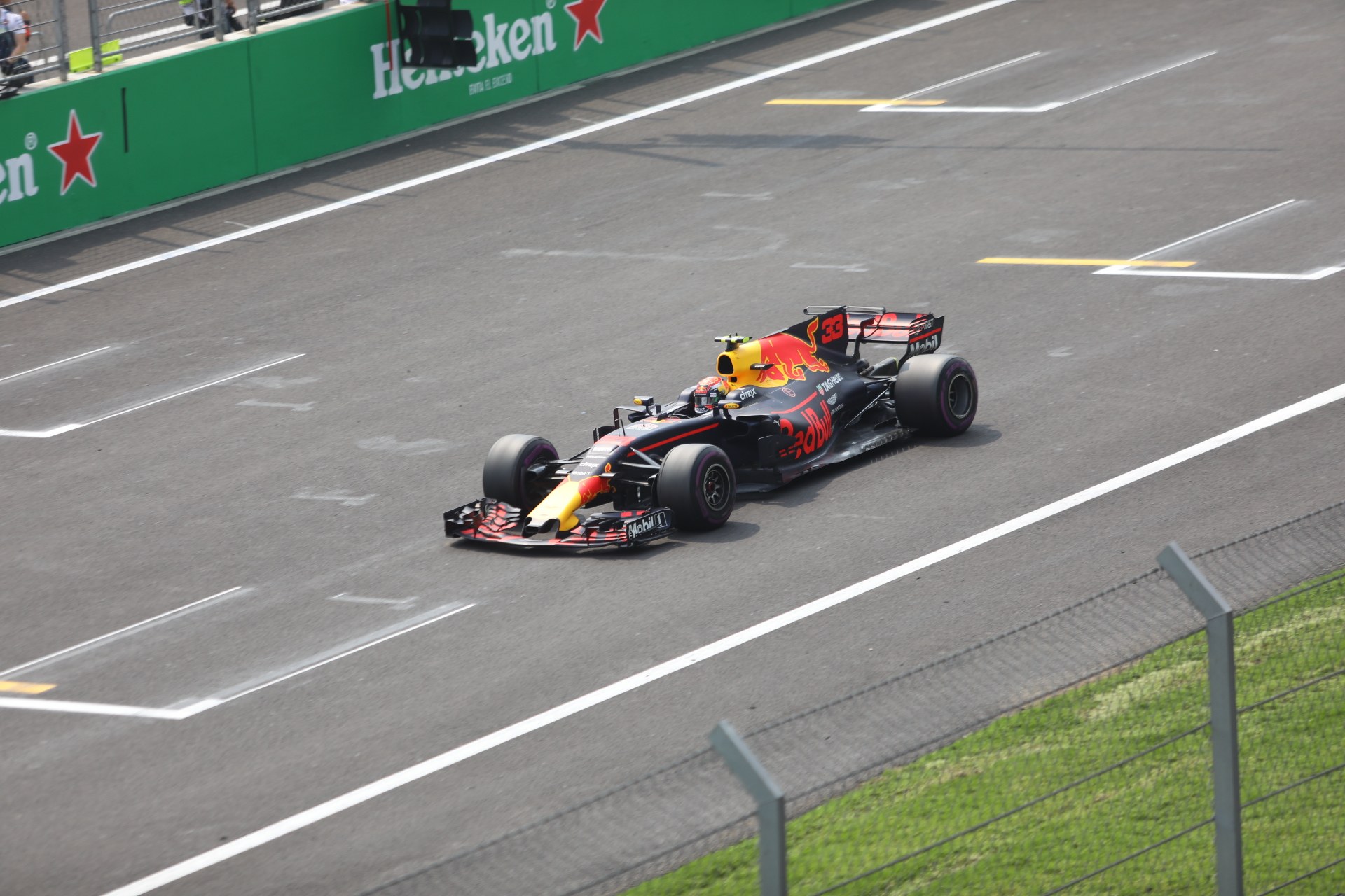 Red Bull Racing car on a race track with a fence and grass to the right, background includes people, railings, sponsor boards, and a traffic signal.