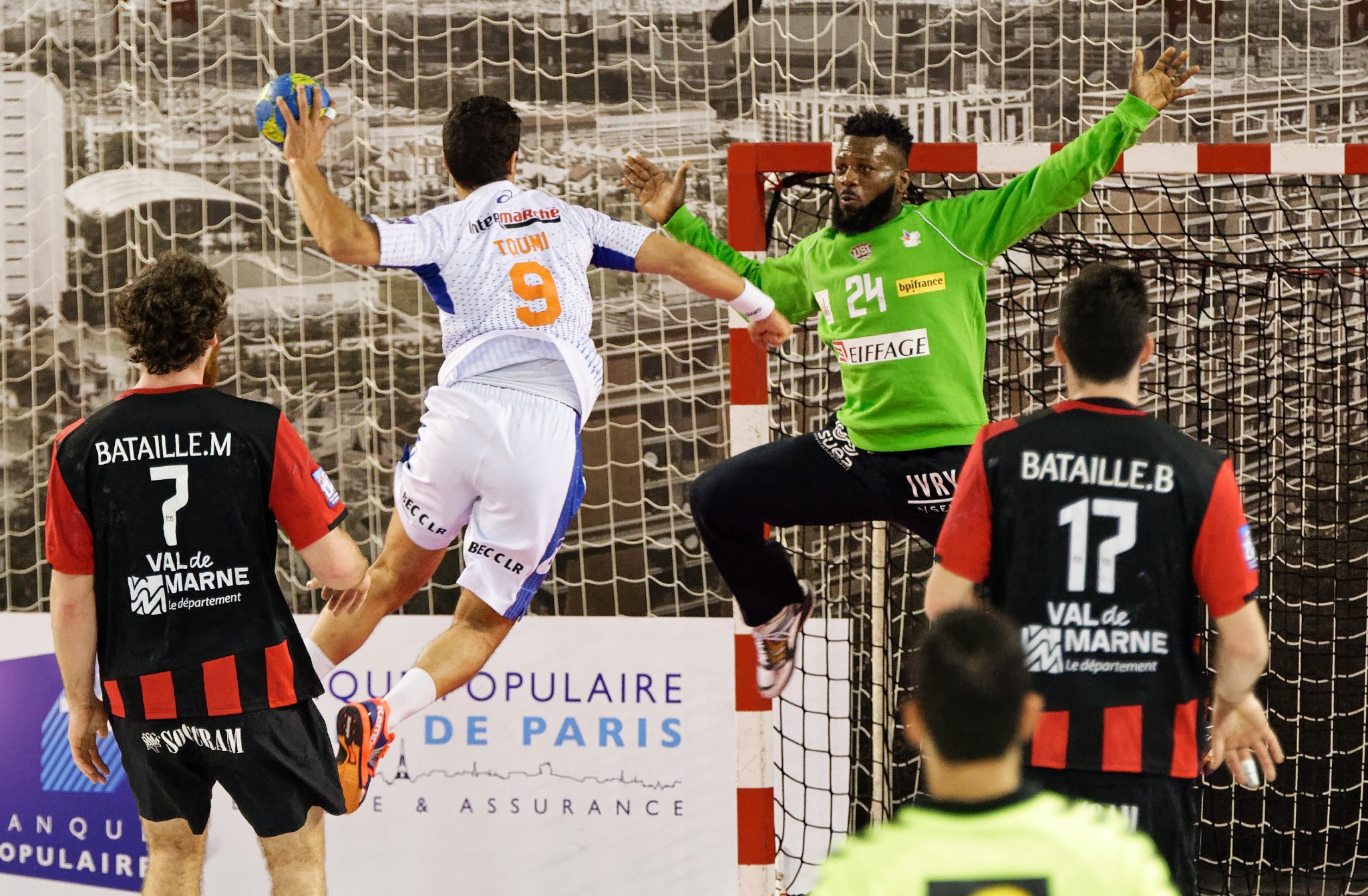 Eine Gruppe von Männern beim Handballspielen, einer springt und hält den Ball, mit einem Torpfosten und einem Banner 'Ligue 1 - Bordeaux vs Marseille' im Hintergrund.