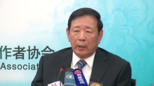 A suited man sits at a table with microphones, speaking during a press event with a banner in the background.