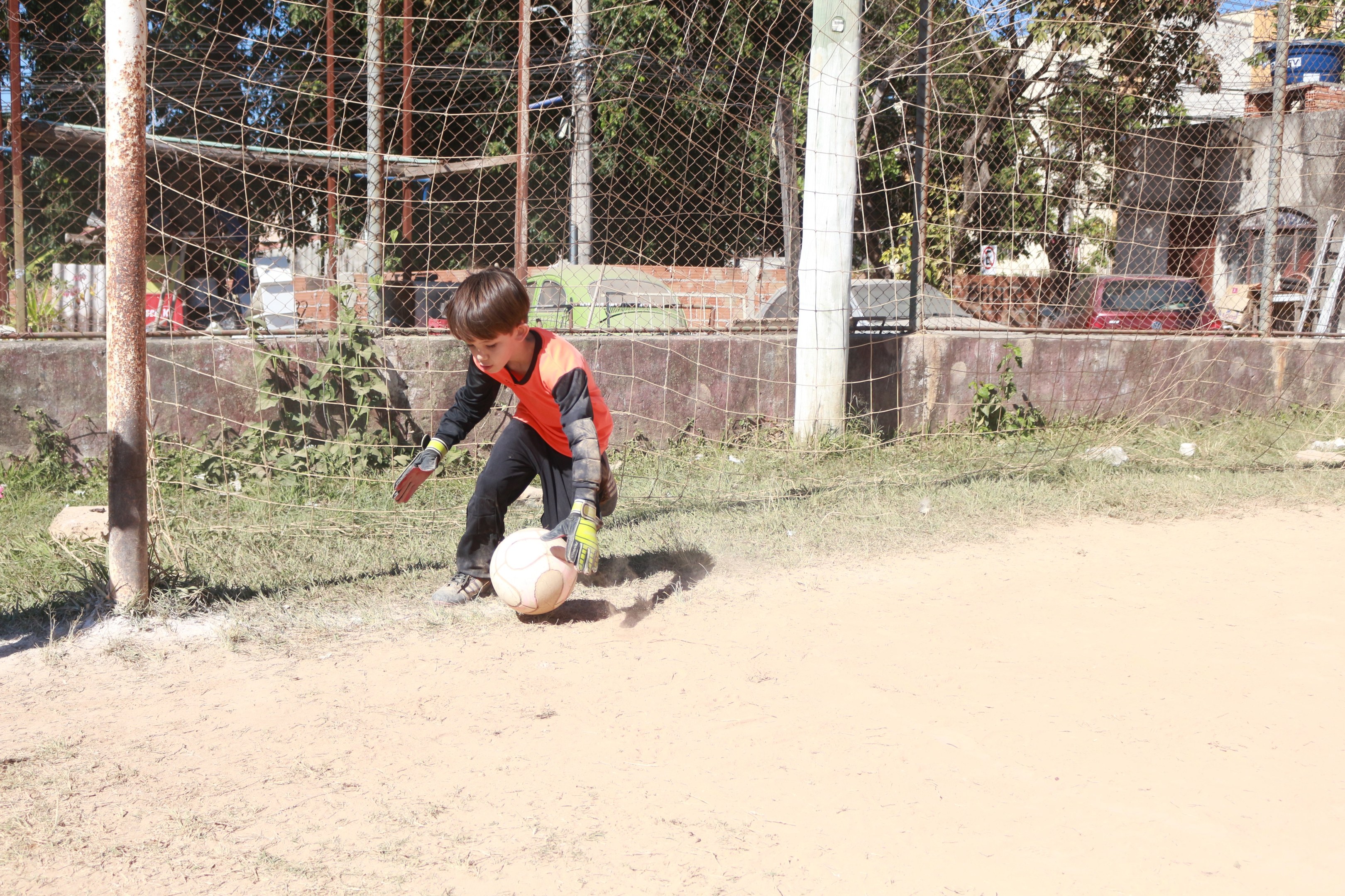 Ein junger Junge spielt Fütball auf einem Schotterfeld mit Gras, Pflanzen, Pfölen, einem Zaun, einer Wand, Bäumen, Fahrzeugen, Gebäuden und einem bewölktem Himmel, trägt Schuhe und ist aktiv im Spiel.