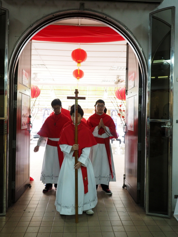 Menschen gehen, während sie ein Kreuz halten, in einer Kathedrale mit Türen links und rechts und Blumen und dekorativen Elementen im Hintergrund.