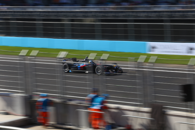 Ein Formelwagen fährt auf einer Rennstrecke an einer Menge Zuschauer vorbei, Gras auf dem Boden und Schilder im Hintergrund.