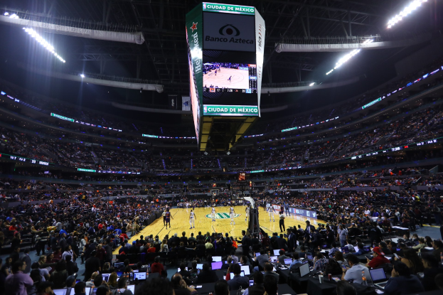 Ein Basketballspiel in einer großen Arena mit Zuschauern auf Sitzen und Stehplätzen, Tischen mit Laptops und Flaschen im Vordergrund und einem Basketballkorb, Anzeigebildschirm und Deckenlampen im Hintergrund.