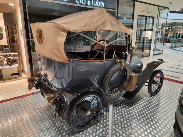 Ein altes Auto in einem Museum ausgestellt, umgeben von Schutzseilen, mit Glaswänden im Hintergrund, die Puppen in verschiedenen Outfits, Lichtern und anderen Gegenst├Ąnden enthalten.