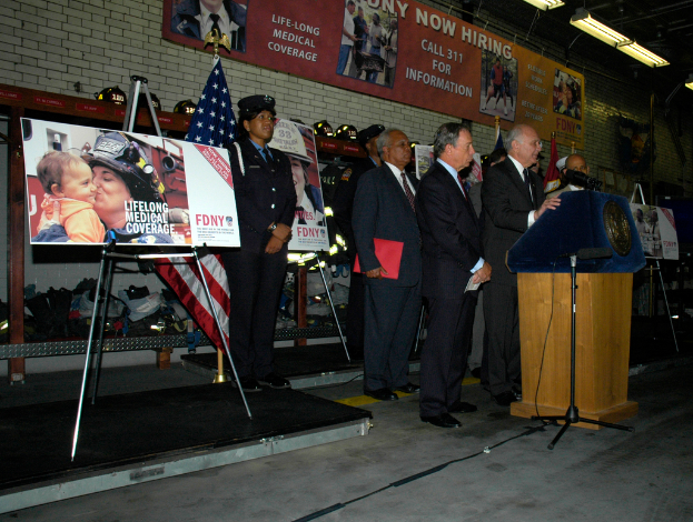 Eine Gruppe von Menschen steht in der Nähe eines Podiums mit Mikrofonen, umgeben von Schildern auf Ständern, Fahnen und Schildern an der Wand, alles unter hellen Lichtern.