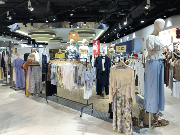 Ein Bekleidungsgeschäft mit Kleiderstangen, Schaufensterpuppen in Outfits, beleuchteter Decke und Informationsschildern mit Text, die einen Mode-Outlet anzeigen.