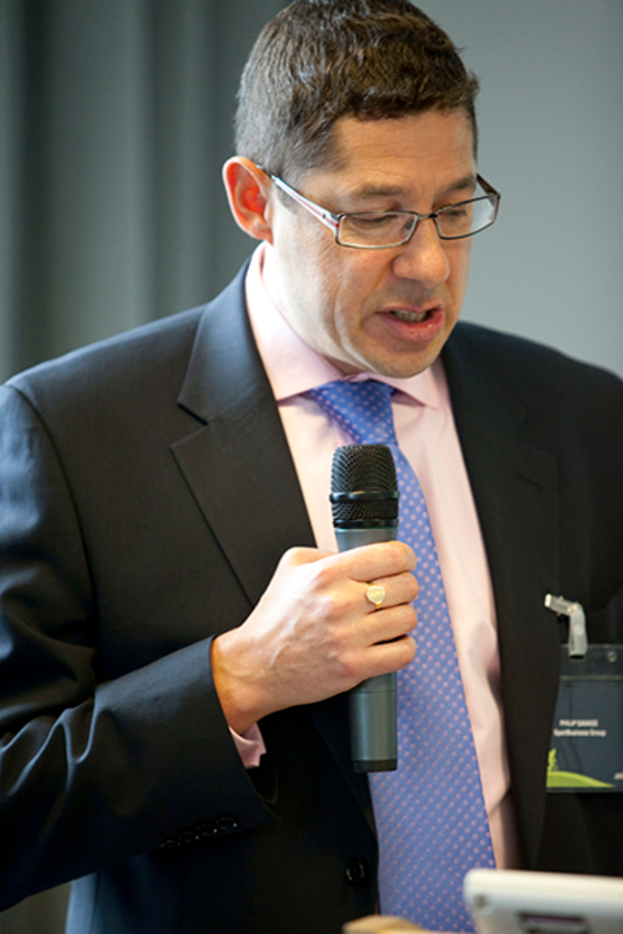 Ein Mann in Anzug, Hemd und Krawatte mit Brille hält ein Mikrofon und spricht, mit einem weißen Abzeichen an seinem Hemd und einem Vorhang im Hintergrund.