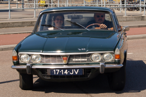 Ein Mann und eine Frau sitzen im Fahrersitz eines grünen Autos auf einer Straße, mit einem Gehweg auf einer Seite und einem Geländer auf der anderen, während Fahrräder, Pfosten, Bretter und Gebäude im Hintergrund zu sehen sind.
