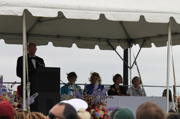 Eine Gruppe von Menschen sitzt an einem Tisch mit Blumen und Flaschen, eine Person steht in der Nähe eines Podiums mit einem Mikrofon und ein Zelt mit Stangen im Hintergrund.
