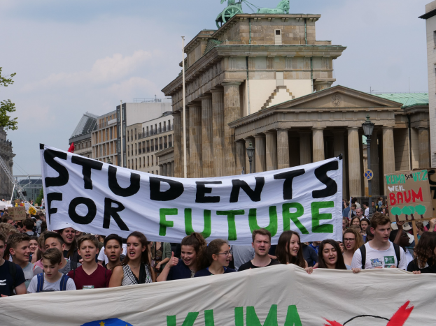 Eine Gruppe von Sch├╝lerinnen und Sch├╝lern marschiert in Berlin, mit einem bunt bemalten Banner, auf dem "Students for Future" steht, vor dem Hintergrund von Geb├Ąuden, B├Ąumen und Himmel.