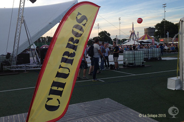 Eine lebhafte Karnevalsszene mit Zelten, einem zentralen Mast, Menschen, Bäumen, Strommasten, Straßenbahnen auf der Straße und einem eisernen Stand auf der linken Seite unter einem klaren Himmel.