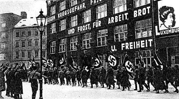 Schwarzes und wei├čes Foto einer Gruppe, die mit Fahnen und Transparenten die Stra├če entlangmarschiert, mit einem Lichtmast links und einer Tafel mit der Aufschrift "Deutsche Nationalsozialistische Arbeiterpartei in Berlin" rechts.