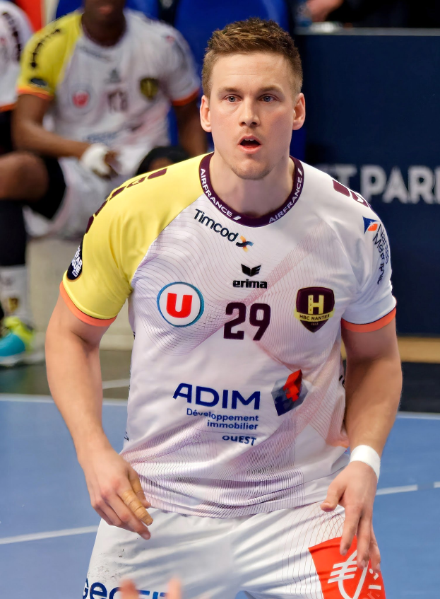Ein Mann in einem weißen Hemd und gelben Shorts spielt Handball, hält ein Objekt und hat einen entschlossenen Gesichtsausdruck, vor einer Tafel mit der Aufschrift "Ligue 1 - Futsal - Olympique de Marseille" und einigen im Hintergrund sitzenden Personen.