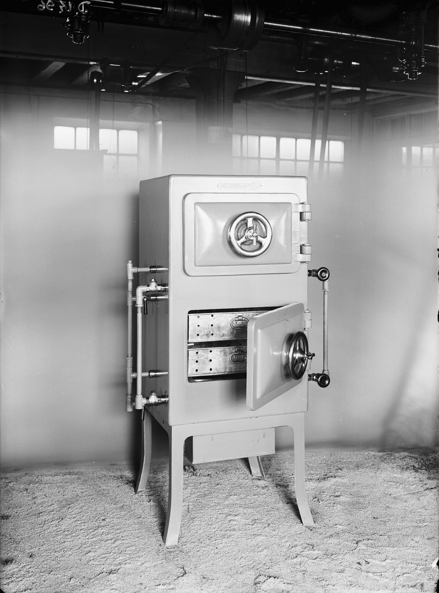 Schwarzes und weißes Foto eines altmodischen Safes in einem Raum mit einer Wand, Fenstern und an der Wand entlanglaufenden Rohren.