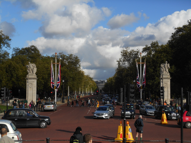 Ein Denkmal steht auf einer Straße mit wenigen fahrenden Autos und Fußgängern, umgeben von Ampeln, Pfählen und Fahnen, mit Bäumen und Gebäuden im Hintergrund unter einem bewölkten Himmel.