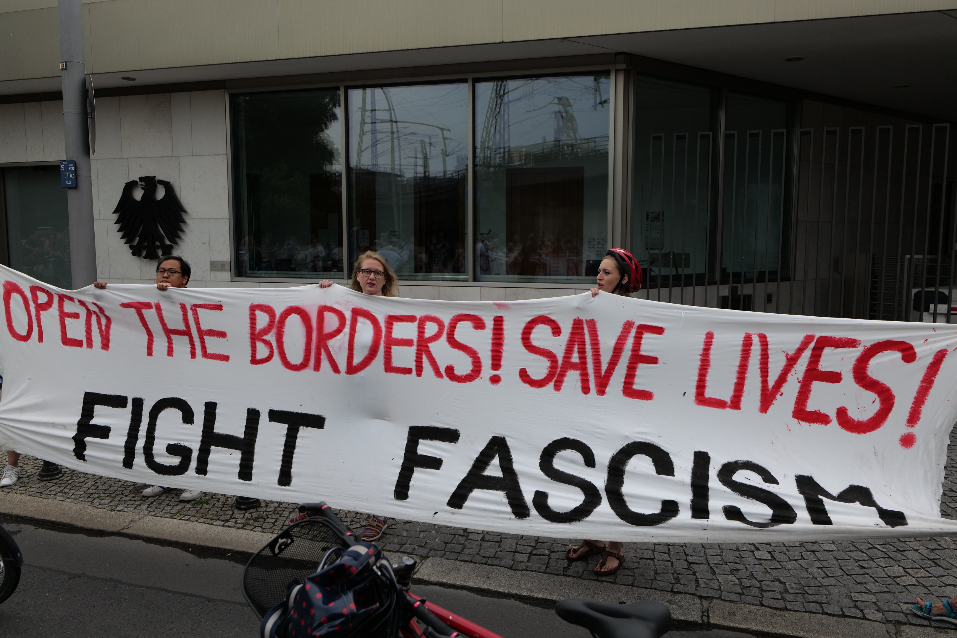 Gruppe von Menschen mit einem Banner "Öffne die Grenzen, rette Leben, bekämpfe Faschismus" vor einem Gebäude mit Glasfenstern und einem Mast, mit einem Fahrrad und einer Tasche auf der Straße.