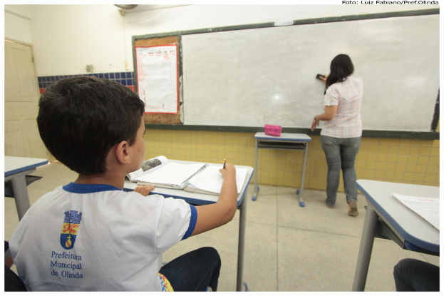 Ein Junge sitzt an einem Tisch und schreibt in ein Buch, während eine Frau das Whiteboard mit einem Putzlappen in einer Klasse mit mehreren Tischen, einem haltend eine Box, und einem Plakat an der Seite putzt.