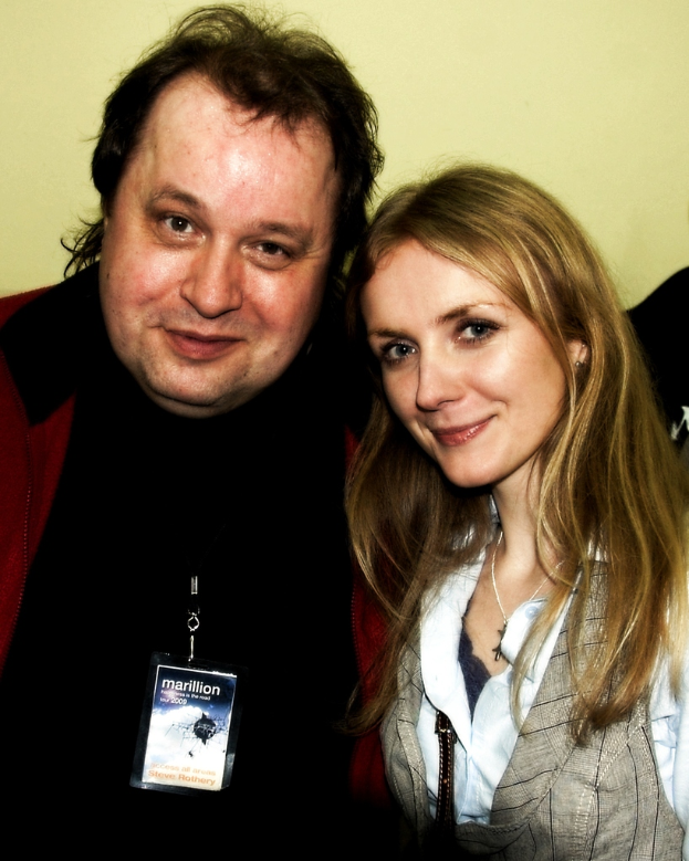 Ein Mann und eine Frau l├Ącheln, mit einem Abzeichen sichtbar, vor einem hellgelben Hintergrund.