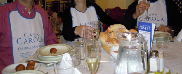 Menschen sitzen an Tischen mit Tellern, Geschirr, Besteck, Brotkörben, Glastumbler und einer Werbung.