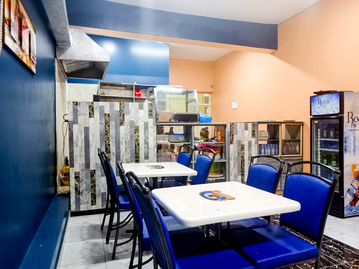 A restaurant with blue chairs and tables, a refrigerator on the right, a framed photo on the left wall, and ceiling lights, located in the Best Western Plus Kochi Airport Hotel.