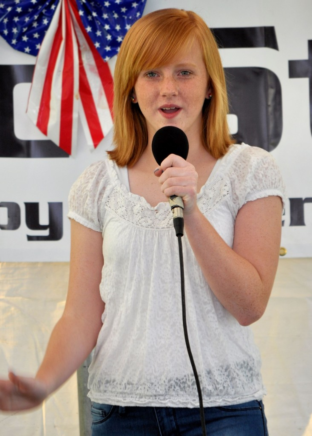 Eine Frau steht an einem Podium, spricht in ein Mikrofon, mit einer USA-Flagge und einem Banner dahinter.