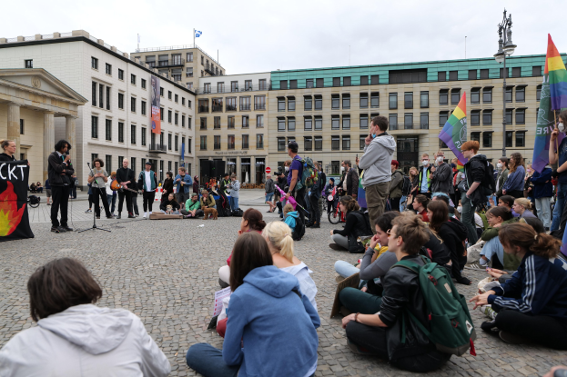 Eine Gruppe von Menschen sitzt auf dem Boden vor einer Menge, die Fahnen und Plakate schwingt, mit einer Person, die in ein Mikrofon spricht, einer Statue und Gebäuden im Hintergrund bei einer Anti-Schwulen-Demo in Berlin.