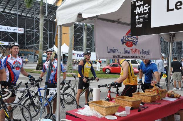 Eine Straßenszene mit Menschen in der Nähe von Fahrrädern, einem Geschäft mit Gegenständen auf einem Tisch, Bäumen, Zelten, an einer Hütte befestigten Schildern, Fahrzeugen und Gras.