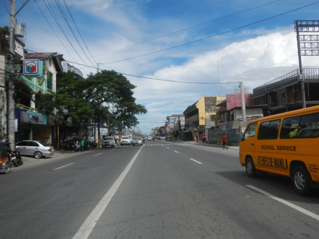 Ein gelber Schulbus fährt eine Straße mit hohen Gebäuden, Menschen auf dem Bürgersteig, Bäumen, Strommasten mit Drähten und Schildern entlang, unter einem bewölkten Himmel.