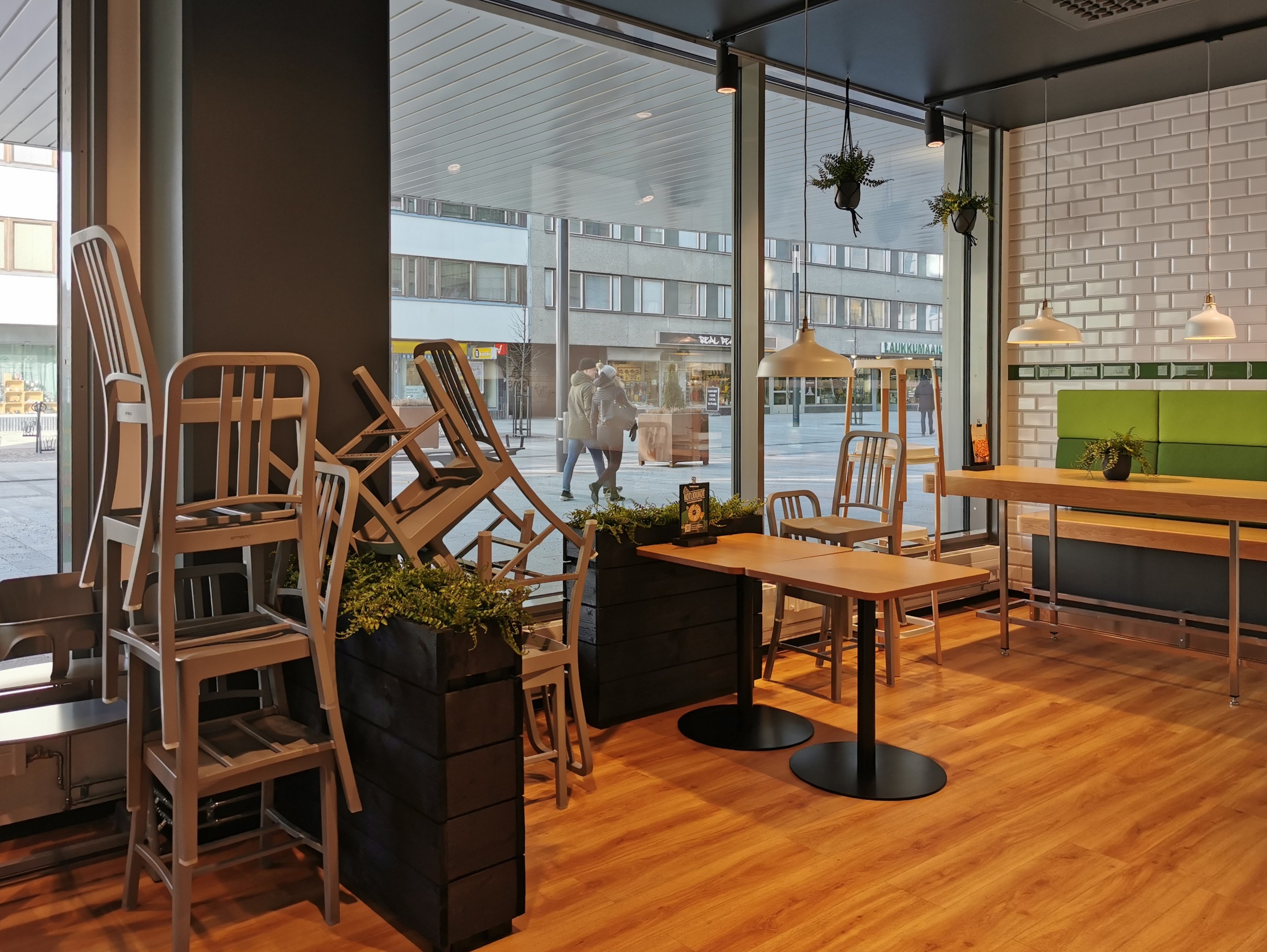 Ein Restaurant-Innenraum mit Holztischen und -stühlen in der Nähe eines großen Fensters, das eine städtische Straßenszene in Berlin zeigt, mit Passanten, Gebäuden und städtischen Elementen.