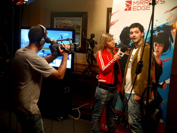 Ein Mann mit einer Videokamera und Headset filmt ein Interview zwischen einem Mann und einer Frau, mit einem Fernseher und einem Foto im Hintergrund.