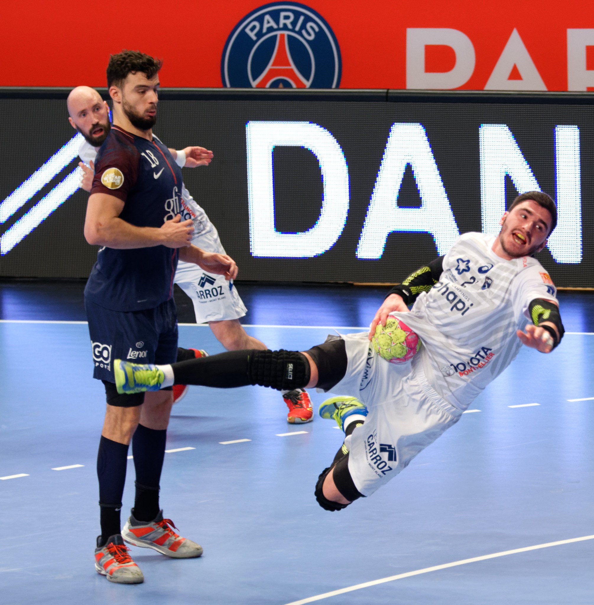 A group of men playing handball on a court, with one holding a ball, and a Paris Saint-Germain futsal team victory board in the background.