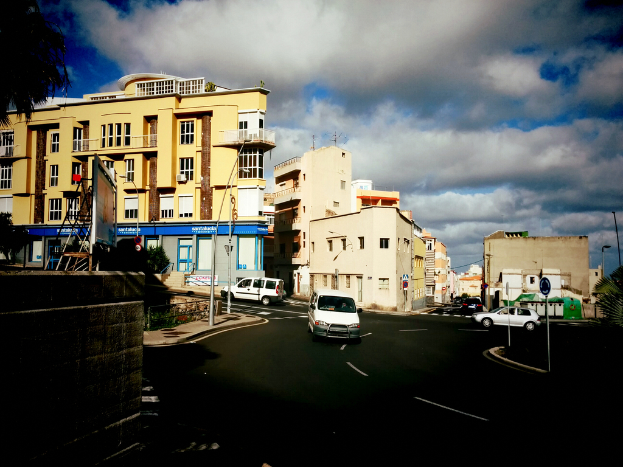Eine stadtstraßenszene mit Fahrzeugen, Polen, Pflanzen, Schildern, Straßenlaternen im Vordergrund, Gebäuden, Autos, Türen, Fenstern und einem Baum in der Mitte, unter einem teilweise bewölkten, sonnigen Himmel.