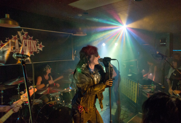 Eine Frau in einer schwarzen Jacke singt in ein Mikrofon auf der Bühne, mit einem Mann in Sonnenbrille und Stirnbinde, der hinter ihr ein Instrument spielt, und einem Plakat mit 'LA Guns' im Hintergrund.