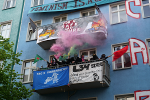 Eine Gruppe von Menschen auf einem Balkon mit Fahnen und Spruchbändern, mit einem Baum links und Rauch in der Luft, während einer Demonstration.