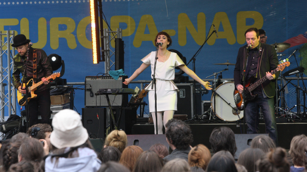 Eine Frau in einem weißen Kleid singt auf der Bühne eines Konzerts, flankiert von zwei Gitarristen, mit Publikum und Lautsprechern im Hintergrund.