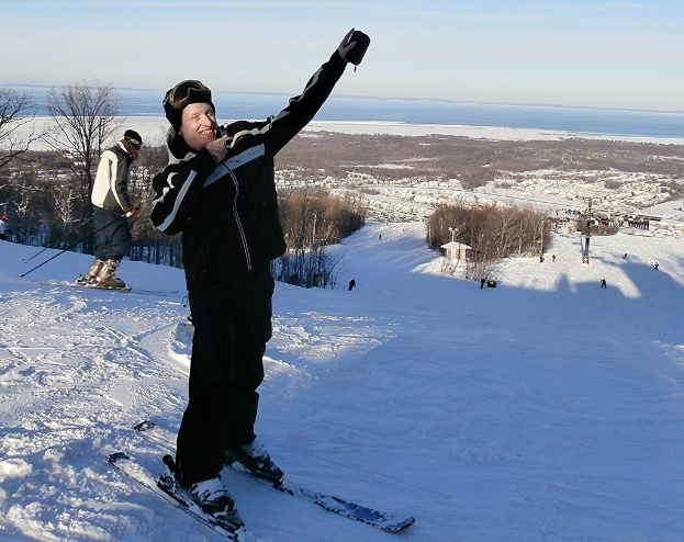 Eine Person auf einem Snowboard fährt eine schneebedeckte Piste mit Bäumen, Menschen, einem Pfahl, Wasser und Himmel im Hintergrund hinunter.