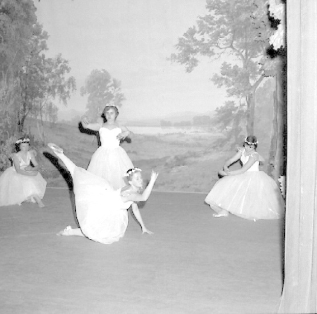 Vier junge Ballerinen in weißen Kleidern tanzen auf einer Bühne mit einer gemalten Kulisse aus Bäumen und Himmel, in Schwarz-Weiß.