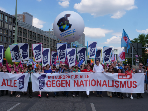 A group of people walking down a street holding a banner reading "Ein Europa für alle Gegen Nationalismus" and flags, with buildings, trees, and a cloudy sky in the background.
