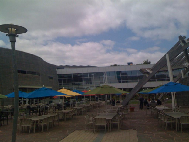 Eine Resort-Szene mit Stühlen, Tischen, Sonnenschirmen und einer Lampe im Vordergrund, Bäume und Gebäude in der Mitte, unter einem bewölkten Himmel.