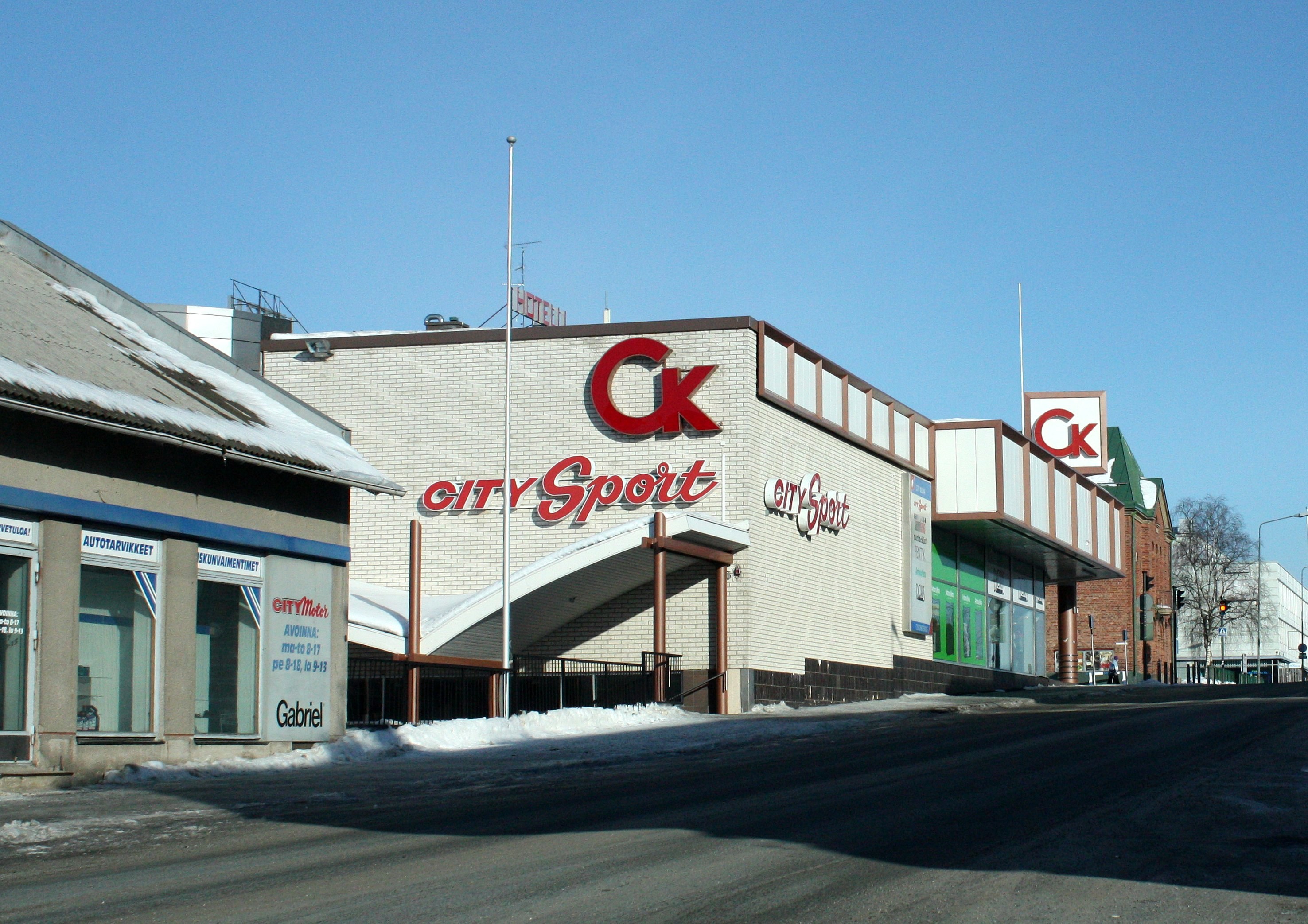 Ein Sportgeschäft in einer Stadt an einer Straßenecke mit umliegenden Gebäuden, Straßenlaternen, Bäumen und Schnee unter einem sichtbaren Himmel.
