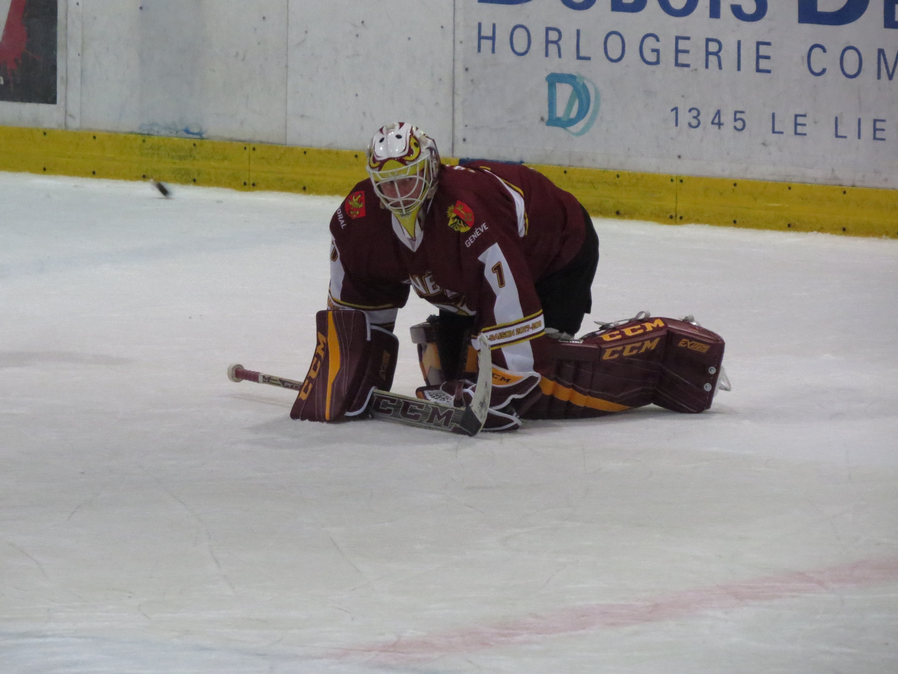 Eishockeyspieler in rot-gelber Uniform bei einem Save auf dem Eis, mit Helm, Handschuhen und Knieschonern, haltend einen Stock, mit einer Wand und Text im Hintergrund.