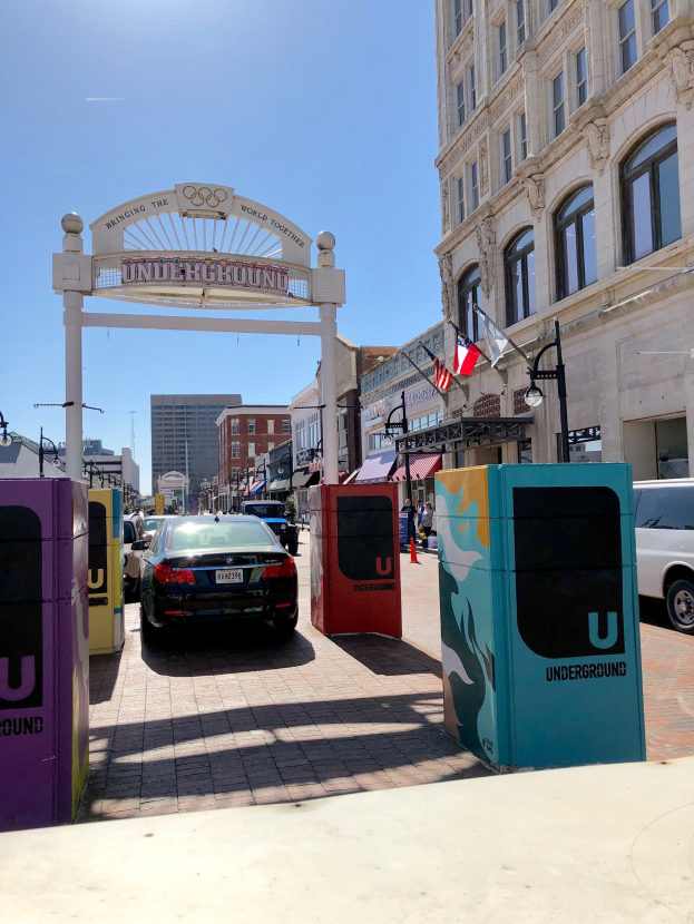 Stadtstraße mit parkenden Autos, ein dekorativer Bogen, Gebäude, Laternen, Fahnenmasten, ein Müllbehälter im Vordergrund und Himmel im Hintergrund.