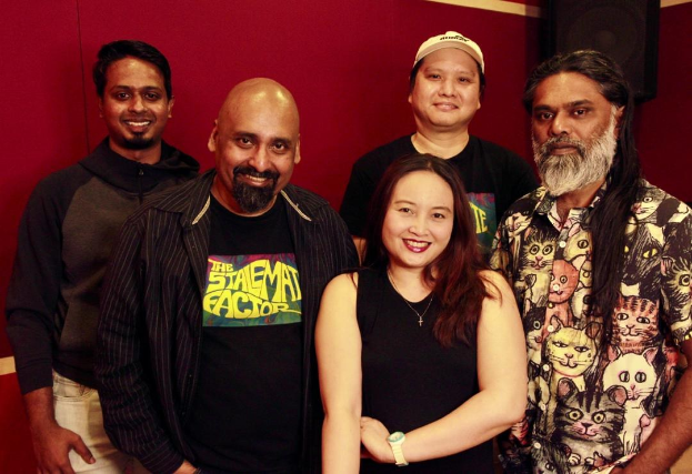 Eine Gruppe von Menschen, die zusammen in einem Aufnahmestudio stehen, lächeln und locker gekleidet sind, mit ihren Armen umeinander gelegt in einer fröhlichen Atmosphäre.